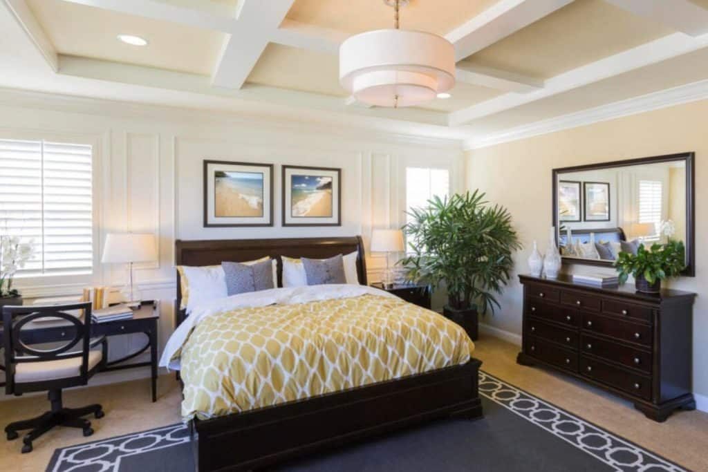 Modern bedroom with a king-sized bed, beige armchair, dark grey walls, wooden floor, and a large window overlooking trees.