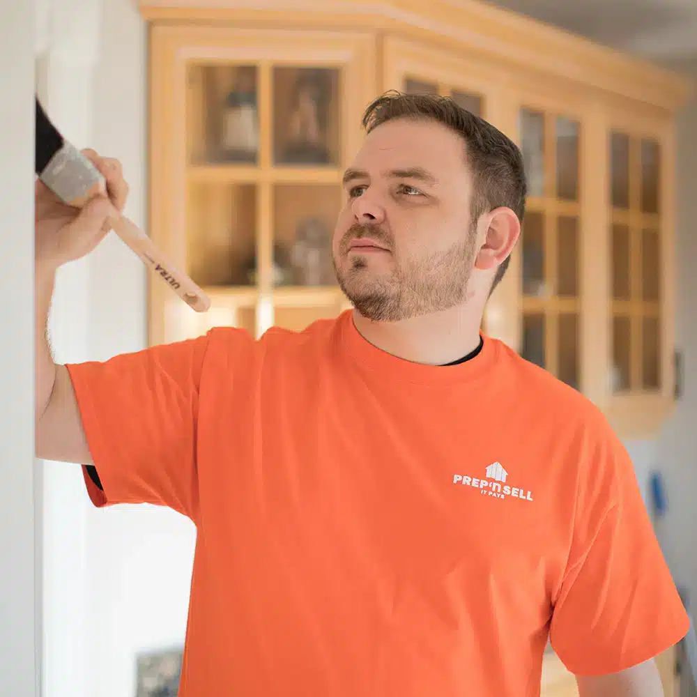 Prep'n Sell team member in a branded orange shirt painting interior trim during a pre-sale home preparation