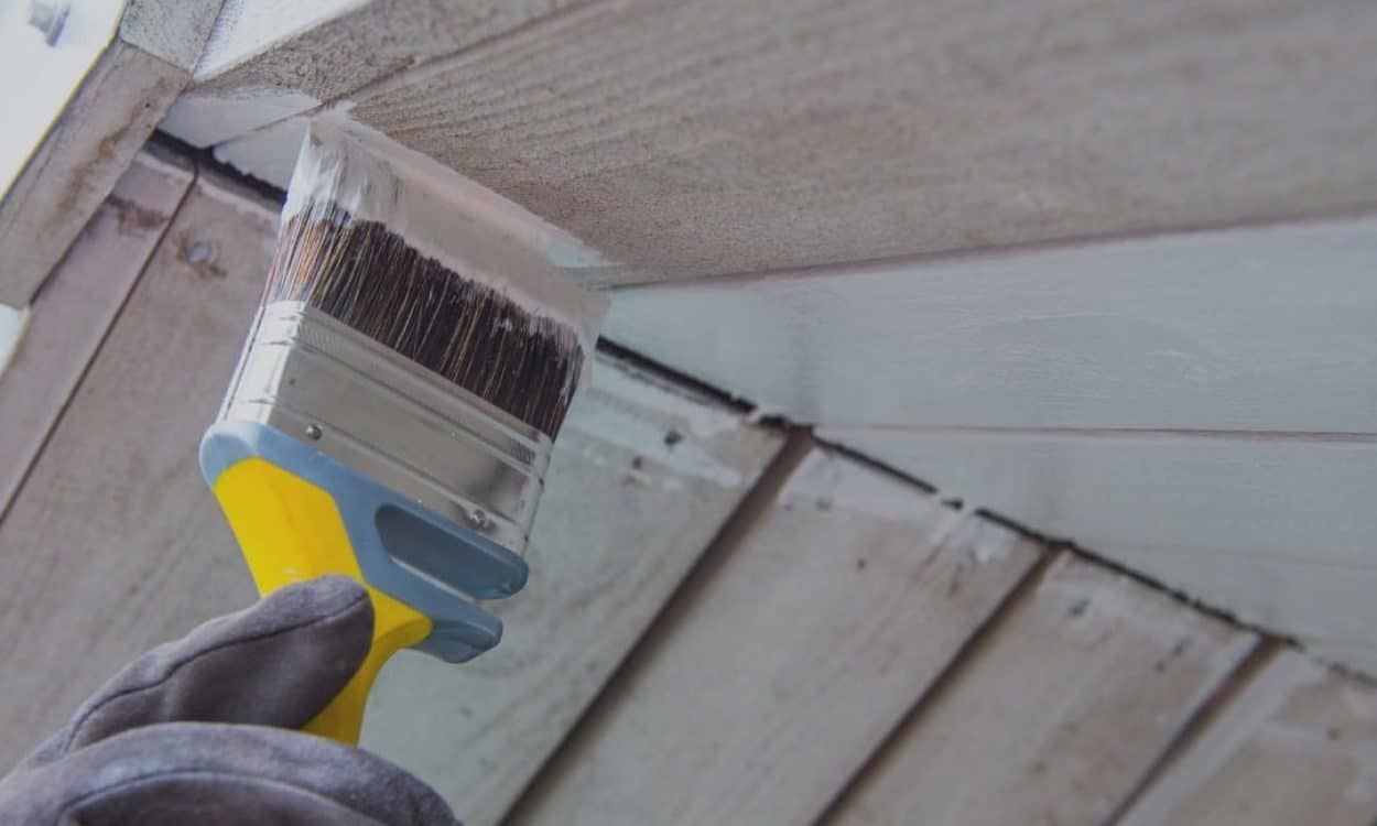 Close-up of a hand holding a paintbrush applying white paint to house siding.